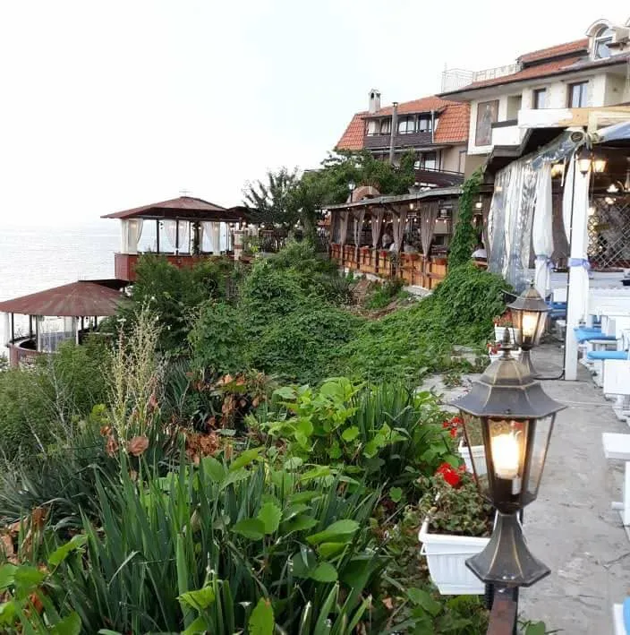 Położenie restauracji Breeze nad brzegiem na Starym Mieście w Nesebyrze: tarasowe drewniane pomosty i altany nad wodą, gęsta zieleń, kuta latarnia i rozciągające się w lewo Morze Czarne.