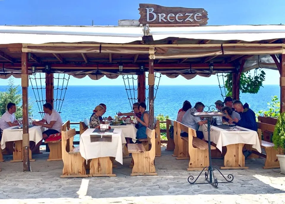 The terrace of Restaurant Breeze from outside: a carved wooden Breeze sign over a timber pergola with canvas roof, pine benches and tables laid with white cloths, guests eating, and the Black Sea directly behind.