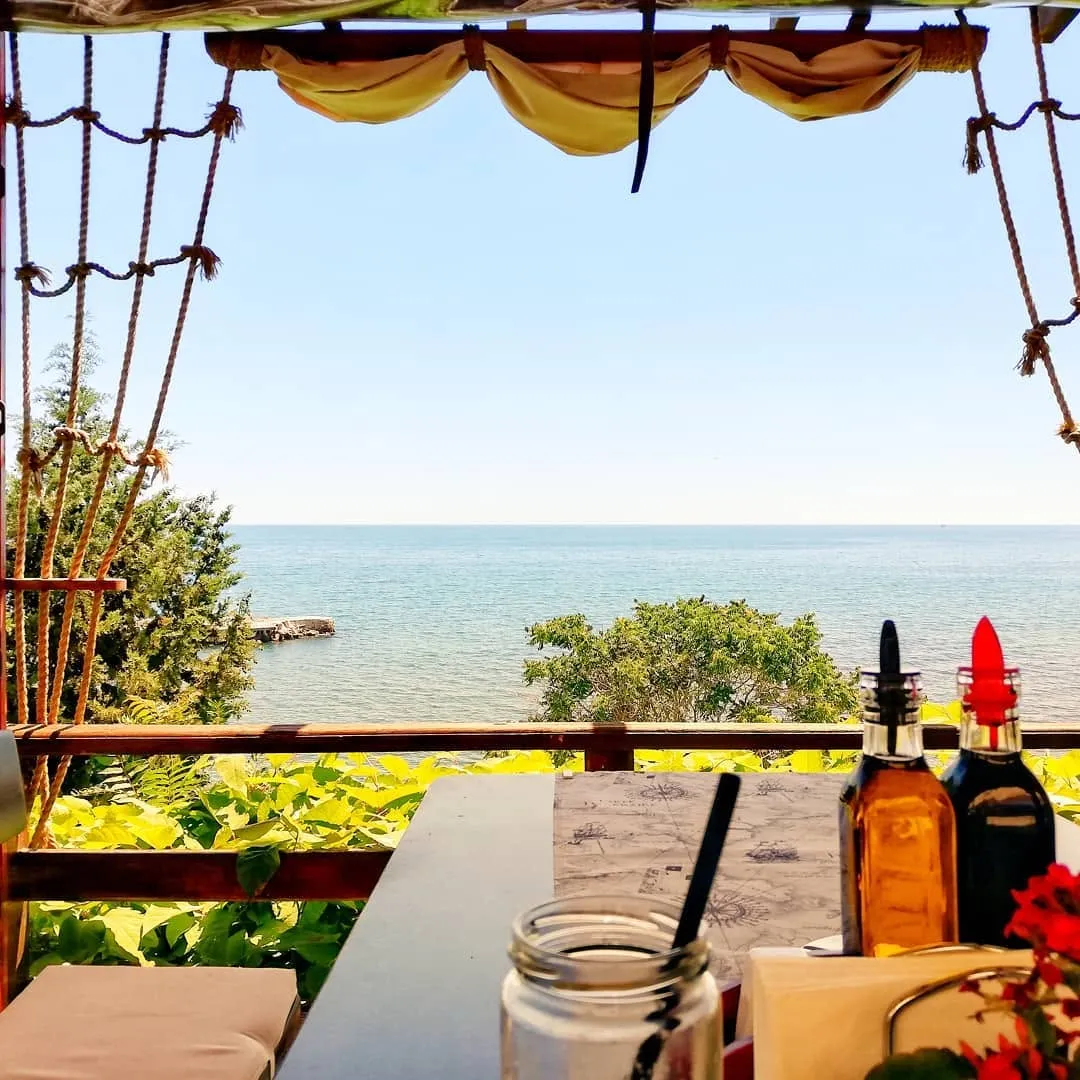 The view from a table on the terrace at Restaurant Breeze: the open Black Sea beyond a wooden rail, framed by rope rigging and a canvas awning, with oil and vinegar bottles on the table.